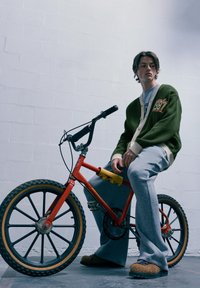 Bicicletta rossa con ruote nere e pneumatici in gomma; modello in cardigan oliva e pantaloni grigi. Calzature marroni con texture, sfondo grigio.