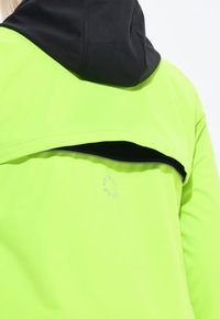 Henkilö, joka on pukeutunut neonvihreään takkiin, jossa on musta huppu ja pyöreä heijastava logo yläselässä, tuuletettu verkkomateriaalilla alla.