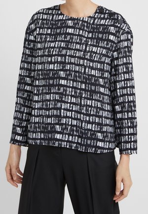 Femme portant un haut à manches longues à motif quadrillé noir et blanc avec des boutons sur l'épaule, associé à un pantalon noir ample.