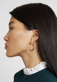 Gouden oorring met een golvend ontwerp, gedragen aan het oor van een model. Het model heeft donker haar en draagt een donkergroene geribbelde trui met een witte gerimpelde kraag.