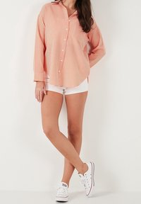 Chemise à manches longues à rayures orange et blanches avec une fermeture boutonnée sur le devant, associée à un short blanc et des baskets basses blanches. Texture de tissu lisse.