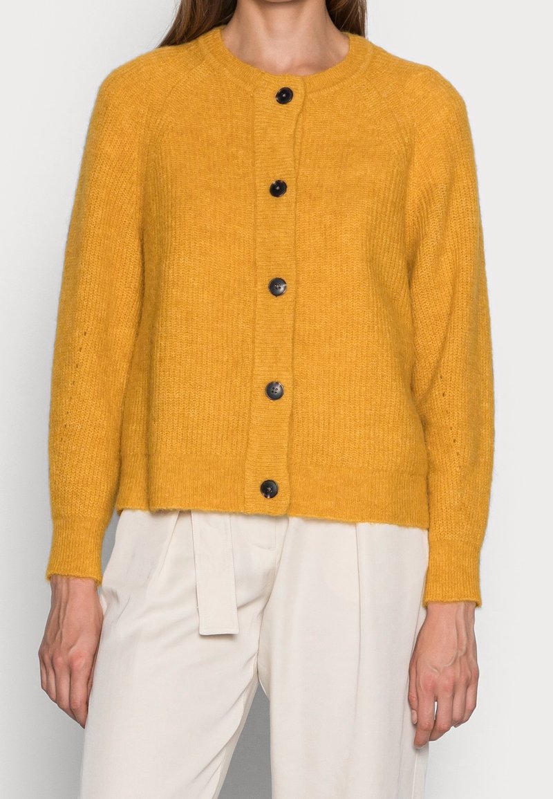 Senapsgul kofta med rund hals, ribbad textur och svarta knappar. Har långa ärmar och en kortad nederkant.