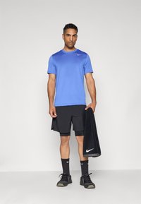 Blå kortærmet træningstrøje, sorte shorts, sorte sokker og sneakers. Holder et sort håndklæde. Nike-logo på trøjen og håndklædet.