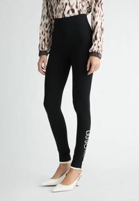 Mallas negras con una cintura alta, que presentan un detalle del logo en el lateral, combinadas con zapatos de punta blanca con tirantes traseros. Tejido suave y elástico.