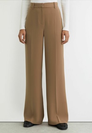 Broek met wijde pijpen van lichtbruin stof. Heeft een platte voorkant, broekriemlussen en een gladde textuur. Draagt zwarte schoenen.