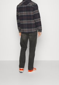 Man wearing black and gray checkered jacket, faded black jeans, and bright orange sneakers with white stripes, standing facing away.