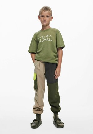 Maglietta verde con grafica bianca, pantaloni cargo beige e verdi con pannelli a blocchi di colore e una tasca verde brillante, abbinati a sandali neri.