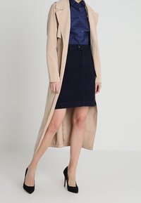 Beige trench coat, tumman sininen nappikauluspaita ja tummat farkkuhameet. Mustat korkokengät viimeistelevät asun. Silkkiset kankaat, klassiset muodot.