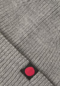 Grau gestrickte Mütze mit gerippter Struktur, einem schwarzen Etikett mit rotem Punktakzent und einem umgeschlagenen Rand.