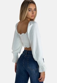 Hvid cropped top med smock-detaljer og puffy lange ærmer, parret med højtaljede blå denimjeans. Glat tekstur, afslappet pasform.