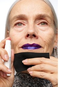 Un tube blanc de produit Wonderskin tenu à côté d'un tissu rectangulaire noir. Le sujet porte un rouge à lèvres violet brillant et présente une texture de peau visible.