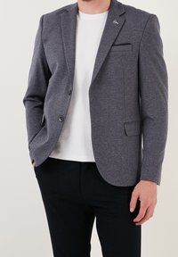 Mężczyzna w szarym marynarce, białej koszuli i czarnych spodniach, stoi z jedną ręką w kieszeni na jednolitym tle.