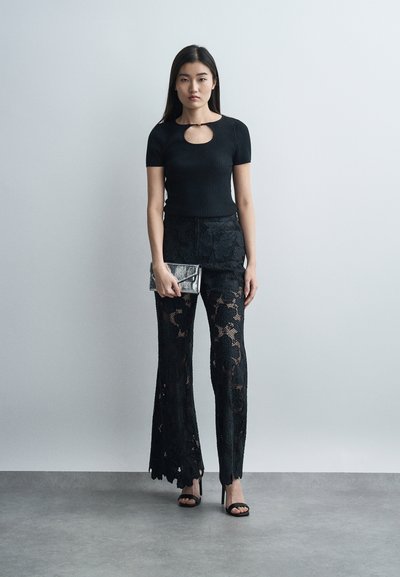 Γυναίκα στέκεται μπροστά σε απλό γκρι τοίχο, φοράει μαύρο κοντομάνικο τοπ με άνοιγμα στο στήθος, μαύρο δαντελένιο παντελόνι, μαύρες γόβες, κρατώντας μεταλλικό ασημί τσαντάκι.