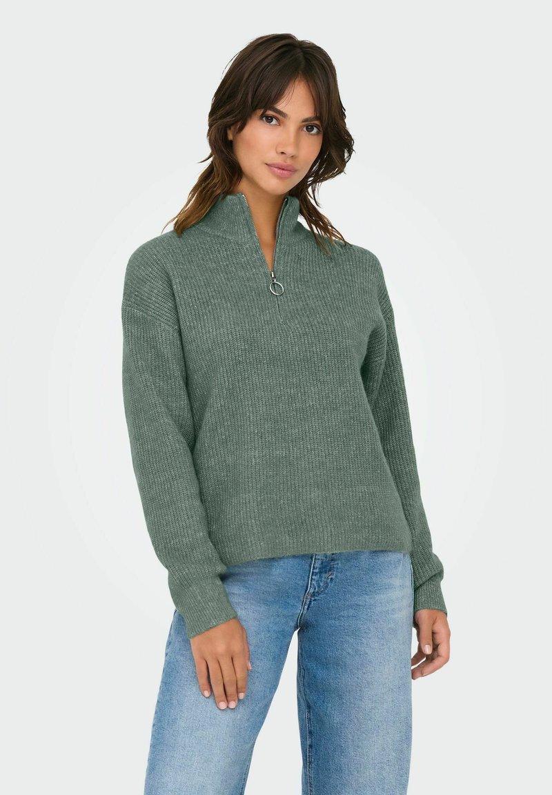 Sweatshirt zippé vert clair en texture côtelée avec un col haut et des manches longues, associé à un jean bleu, présenté sur un fond neutre.