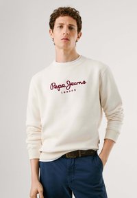 Sudadera blanca con el logotipo bordado en burdeos "Pepe Jeans LONDON", con cuello redondo y puños de canalé, llevada sobre una camiseta blanca, combinada con pantalones de color marino.