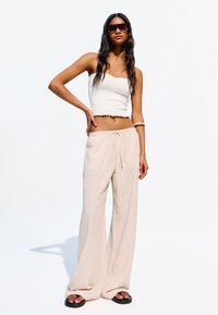 Top court blanc à hem ruffled, associé à un pantalon large beige à rayures et des tongs noires. Modèle portant des lunettes de soleil foncées.
