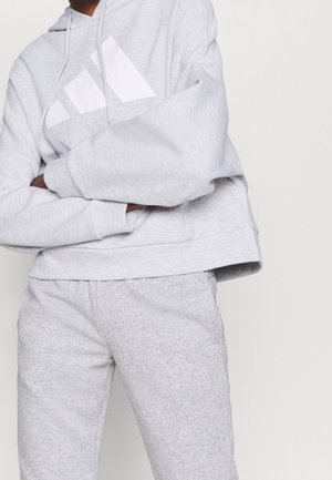 Persoon draagt lichtgrijze Adidas hoodie en bijpassende joggingbroek met gekruiste armen tegen een effen achtergrond.