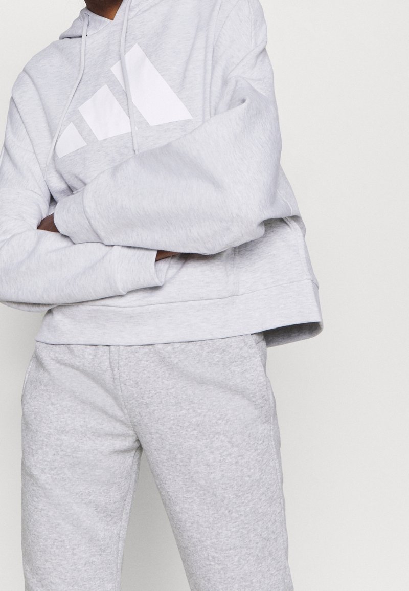 Personne portant un sweat à capuche Adidas gris clair et un pantalon de survêtement assorti, les bras croisés devant un fond uni.