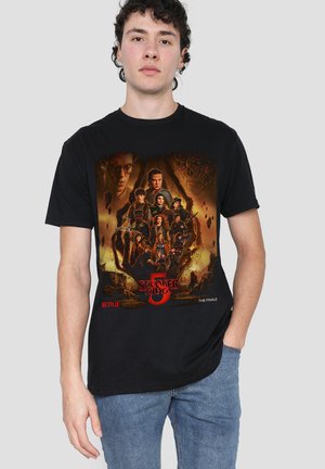 Young man wearing black Stranger Things Season 5 finale T-shirt with character collage and fiery background standing against plain gray backdrop.