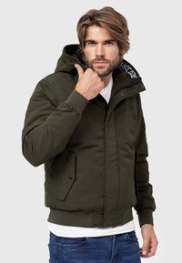 Giubbotto bomber color khaki con cappuccio, zip frontale, bottoni a pressione, polsini elasticizzati e due tasche laterali. La fodera interna presenta un motivo scuro.