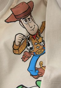 Béžové tričko s potiskem karikaturního kovboje s hnědým kloboukem, kostkovanou košilí a modrými kalhotami. Design obsahuje šerifské odznaky a jemné detaily.