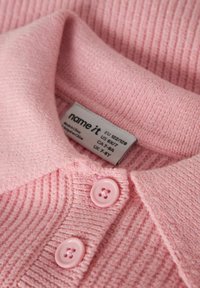 Polo rosa lavorata a maglia con trama a coste, caratterizzata da un colletto morbido e due bottoni rosa. Etichetta di cura visibile con informazioni su taglia e marca.