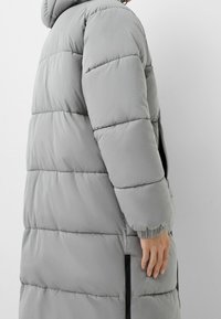 Šedý péřový kabát s horizontálně prošívaným designem, dlouhými rukávy a bočními zipovými kapsami. Obsahuje kapuci a elastické manžety pro tepelnou izolaci.