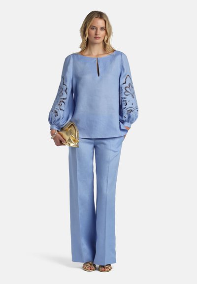 Vrouw die een lichtblauwe blouse met lange mouwen draagt met bloemenuitgesneden details op de mouwen, bijpassende wijde broek, een gouden clutch en gouden sandalen.