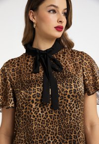 Bluză cu imprimeu leopard dintr-un material maro transparent, având un guler negru cu volănașe și o fundă legată la gât, cu mâneci scurte, evazate.