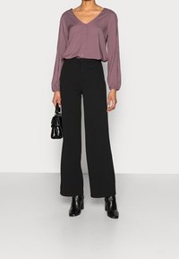 Mujer vistiendo una blusa de botón en color malva con escote en V, pantalones anchos negros, botas negras de tobillo, sosteniendo un pequeño bolso de mano negro.