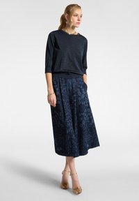 Gonna lunga blu navy con motivo floreale e tasche, abbinata a una maglietta scura e luccicante a fit largo. Sandali con tacco dorati completano l'outfit.