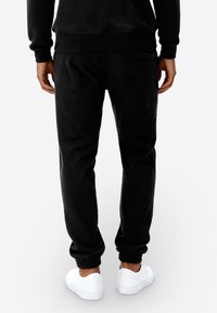 Zwarte joggingbroek van zacht stof, voorzien van een elastische tailleband en boorden, met een achterzak en gedragen met witte sneakers.