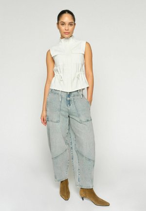 Žena oblečená v bielom tielkovom top s na zips, voľných svetlomodrých džínsoch s veľkými vreckami a hnedých semišových členkových topánkach, stojaca na jednoliatom pozadí.