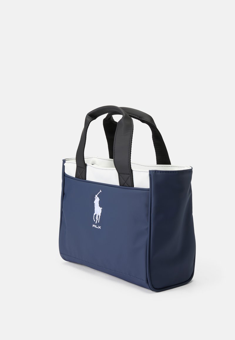 Bolso tipo tote de color azul marino con asas negras, hecho de material suave. Cuenta con un interior blanco y un logotipo bordado en la parte frontal. Diseño compacto.