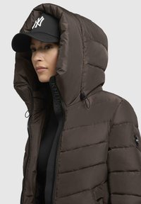 Giubbotto puffer marrone con un ampio cappuccio, chiusura a zip e dettagli elasticizzati. Presenta una toppa con logo nera e un cappellino abbinato nero con emblema bianco.