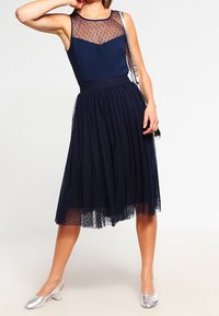 Abito navy con sovrapposizione in rete trasparente a pois sul corpetto, gonna di tulle plissettata e scarpe con tacco a blocco argentate. La borsa a tracolla esalta il look.