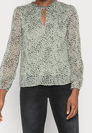 Blouse légère verte clair, transparente avec un motif moucheté noir, décolleté rond avec fermeture à lacer et manches élastiquées. Associée à un jean foncé.