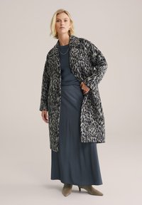 Cappotto maculato in grigio e nero, con colletto a punta e apertura frontale, indossato sopra un top blu scuro e una gonna grigia, con tacchi taupe.