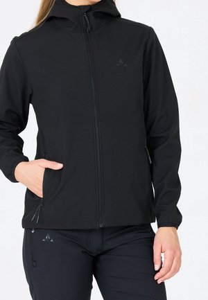 Femme portant une veste noire zippée à capuche et un pantalon noir, les mains dans les poches sur fond blanc.