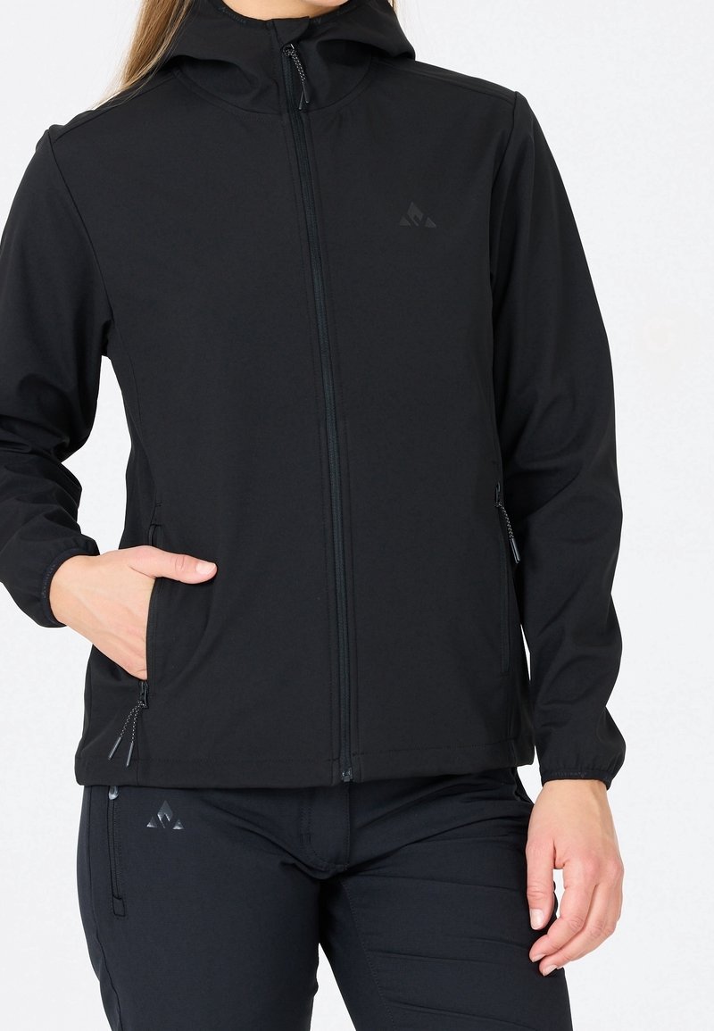 Femme portant une veste noire zippée à capuche et un pantalon noir, les mains dans les poches sur fond blanc.