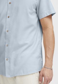 Lichtblauw kort shirt met een soepele textuur, voorzien van houten knopen en een klein zijlabel. Gecombineerd met crème kleurige broek.