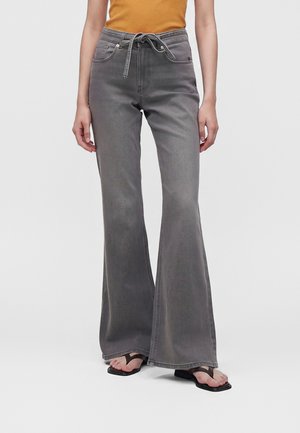 Personne portant un jean flare gris taille haute avec un lien à l’avant, une chemise moutarde et des sandales noires à bout carré avec les orteils visibles.