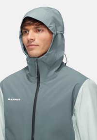 Veste imperméable grise avec capuche, dotée d'un col montant et d'une fermeture éclair à l'avant. Nom de la marque "MAMMUT" en blanc sur le côté gauche. Texture lisse.