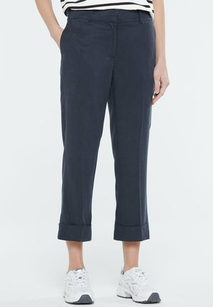 Marineblaue, gekürzte Hose mit Umschlägen, getragen mit weißen Sneakers und einem gestreiften Oberteil, Hände ruhen an den Seiten und in den Taschen.