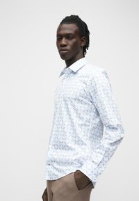 Jeune homme à la peau sombre et aux cheveux tressés, portant une chemise blanche à motif géométrique bleu clair et un pantalon beige, la main dans la poche.