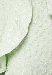 O tecido verde-claro apresenta um design texturizado com padrões florais em branco, criando um contraste subtil. Textura suave, detalhes delicados por todo o lado.