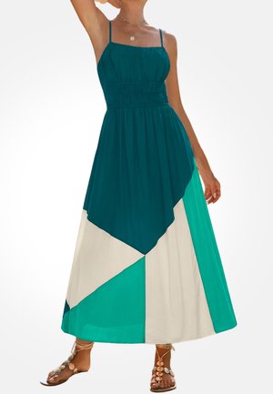 Vrouw draagt een mouwloze maxi-jurk met teal, wit en turquoise geometrische panelen en sandalen met decoratieve schelpen.