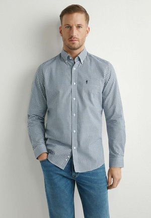 Mężczyzna w niebiesko-białej koszuli w kratę i niebieskich dżinsach, stojący na tle jednolitej jasnej ściany, z jedną ręką w kieszeni.