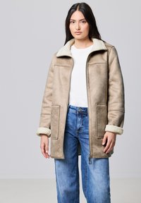 Abrigo de shearling sintético beige con cremallera en el frente, grandes bolsillos y puños esponjosos. Combinado con jeans de corte holgado azul claro y una camisa blanca.