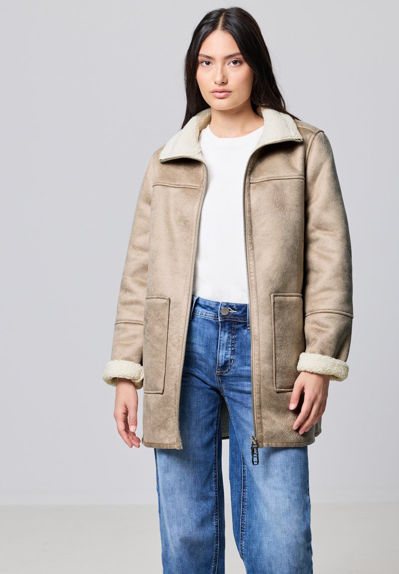 Abrigo de shearling sintético beige con cremallera en el frente, grandes bolsillos y puños esponjosos. Combinado con jeans de corte holgado azul claro y una camisa blanca.